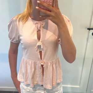Ljusrosa blus med rosetter från Gina Tricot - Supersöt ljusrosa blus från Gina Tricot med puffiga korta ärmar och tre rosetter framtill. Den är lite öppen men fu kan ha ett linne under! 
