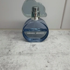 Ariana Grande Cloud parfym 50ml - Säljer en Ariana Grande Cloud parfym i en rund, blå glasflaska med ett unikt lock i form av ett moln. Flaskan har silvriga detaljer och texten 'Ariana Grande' på framsidan. Perfekt för dig som gillar fräscha och söta dofter.