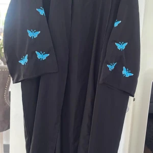 Svart långklänning med broderade fjärilar, Storlek Onesize 36/46 passar! - Två stycken helt oanvända svarta långklänningar med vida ärmar och broderade fjärilar i blått och rosa på ärmarna. Klänningen har en loose passform och är tillverkad i ett lätt, flowy material. Perfekt för dig som vill sticka ut med coola detaljer och färgglada broderier. Styck 170 kr. Klänningarna är Onesize där de passar ifrån storlek 36-46