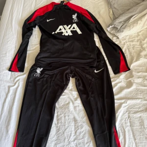 Liverpool Nike träningset svart/röd 24/25 - Liverpool FC träningströja i svart med röda detaljer på axlar och ärmar. Långärmad modell med half zip och vita loggor framtill. Byxorna passar ihop till tröjan. Inget av det är använt och därför i topp skick. Hör av er vid frågor! Man kan köpa bara tröjan eller bara byxorna också. Hör av er! Pris kan diskuteras vid snabb affär. 
