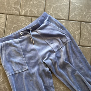 Blå velour mjukisbyxor Juicy Couture - Blåa mjukisbyxor i mjuk velour från Juicy Couture med bred resår och snörning i midjan. Stora fickor fram och bak, varav bakfickan har tryckknappar. Broderad JC-logga på ena fickan. Perfekta för en chill och trendig look.