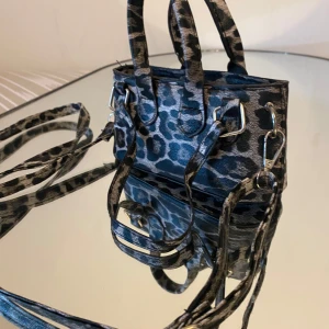 Leopardmönstrad minihandväska - Säljer en liten och trendig handväska med leopardmönster i svart och grått. Väskan har både handtag och lång axelrem, perfekt för att bära crossbody eller i handen. Snygga silvriga metalldetaljer och fyrkantig form. Perfekt accessoar för att lyfta din outfit.