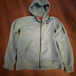 Ljusblå Nike hoodie med dragkedja - Nike hoodie i ljusblått med hel dragkedja och huva. Tröjan har fickor framtill, en extra ficka med dragkedja på ärmen och klassisk Nike-logga på bröstet. Perfekt för chill eller träning, mjukt material och relaxed fit. 