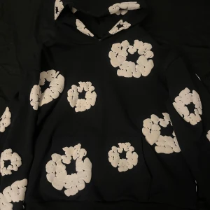 Svart Denim Tears Hoodie - Säljer en svart hoodie från Denim Tears med ett unikt mönster av vita blommor. Hoodien har en klassisk passform med en stor ficka framtill och en bekväm huva. Perfekt för att ge din outfit en cool touch.