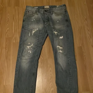 Ljusa Jack and Jones jeans med slitningar - Säljer ett par ljusa jeans med coola slitningar framtill. De har en klassisk femficksdesign och en knappgylf. Perfekta för en avslappnad stil.