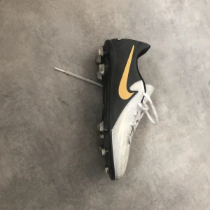 Nike Phantom GT fotbollsskor - Säljer ett par Nike Phantom GT fotbollsskor i svart och vitt med gulddetaljer. Skorna har snörning och en rund tå, perfekt för fotbollsplanen. De har en stilren design med ett mönster på ovansidan för bättre bollkontroll.
