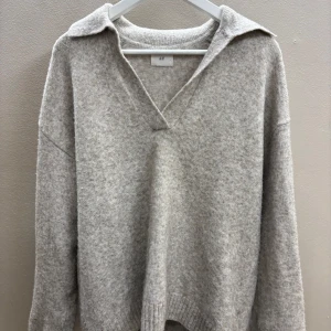 Grå stickad tröja från H&M - Säljer en stilren grå stickad tröja från H&M med v-ringning och krage. Tröjan har långa ärmar och ribbade kanter som ger en klassisk look. Perfekt för kyligare dagar🥶tröjan är ganska lång i både armar och på längden så den är lite oversized! Väldigt fin och inte använd många gånger! 💕💕
