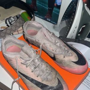 Nike fotbollsskor i beige och rosa - Snygga Nike fotbollsskor i beige med svarta detaljer och rosa inslag. Skorna har snörning och en modern design som passar perfekt för fotbollsplanen. Finns ett litet hål på högra skon men inget som påverkar spelet.