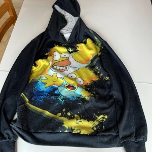 Svart hoodie med tryck - Cool svart hoodie med färgglatt tryck av tecknade figurer på framsidan. Hoodien har en stor ficka framtill och en bekväm huva. Perfekt för en avslappnad stil!