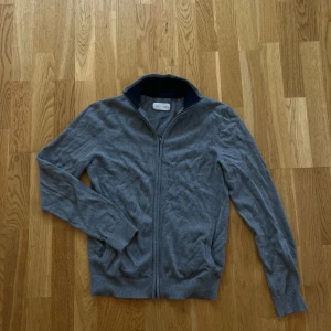 Grå zip kofta - Säljer en stilren grå kofta med dragkedja och ribbad krage. Perfekt för lager-på-lager och passar till många olika outfits. Koftan har långa ärmar och är tillverkad i ett mjukt material.