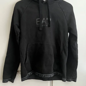 Svart hoodie från Emporio Armani - Snygg svart hoodie från Emporio Armani med logga på bröstet och ribbad kant med märkesdetaljer. Den har en klassisk känguruficka och justerbar dragsko i huvan. Perfekt för en stilren och bekväm look.