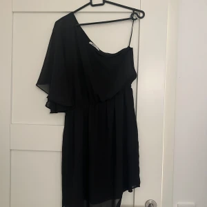 Svart oneshoulder klänning - Snygg svart oneshoulder klänning med en luftig design. Perfekt för en kväll ute eller en speciell tillställning. Den har en elegant asymmetrisk stil med en axelbandslös sida och en tunn axelrem på andra sidan.