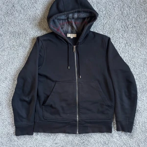 Burberry Hoodie - Säljer en stilren svart hoodie från Burberry med dragkedja och klassiskt rutigt foder i grått och rött. Perfekt för en avslappnad och trendig look. Priset går att diskutera! Hör av er vid frågor och funderingar! MVH Lavish. 