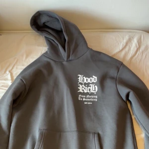 Grå hoodie från Hoodrich - Säljer en grå hoodie från Hoodrich med tryck både fram och bak. Texten 'Hood Rich' pryder framsidan och baksidan har ett stort grafiskt tryck med olika texter. Hoodien har en klassisk känguruficka och en bekväm huva. Perfekt för en avslappnad stil. Storlek M men känns som L!