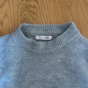 Ljusblå stickad tröja från Design - Säljer en mysig ljusblå stickad tröja från Design by si. Tröjan har en klassisk rund hals och långa ärmar med ribbade muddar vid ärmslut och nederkant. Perfekt för kyliga dagar!