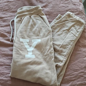 Beige mjukisbyxor från Yale - Säljer ett par bekväma beige mjukisbyxor från Yale med en stor vit bokstav 'Y' på sidan. Byxorna har resår i midjan och snörning för justerbar passform. Perfekta för en avslappnad stil.