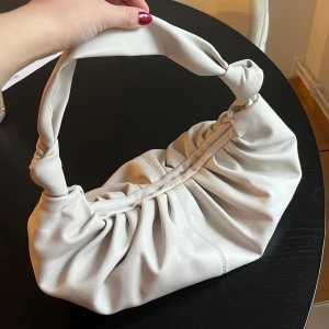 Vit väska med knutdetaljer - Snygg vit axelväska från Gina Tricot med unika knutdetaljer på handtaget. Väskan har en mjuk och rynkad design som ger en trendig look. Perfekt för att lyfta din outfit med stil. Inget innerfack. 