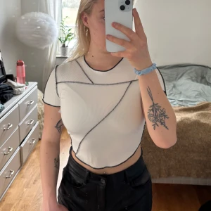 Vit ribbad croppad topp - Säljer en vit ribbad croppad topp med svarta kontrastsömmar. Toppen har korta ärmar och en rund halsringning. Perfekt för en trendig look!