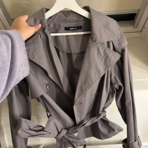 Grå trenchcoat från Gina Tricot - Snygg grå trenchcoat från Gina Tricot med knytbälte i midjan och klassisk krage. Jackan har långa ärmar och knappar framtill. Perfekt för höst och vår!