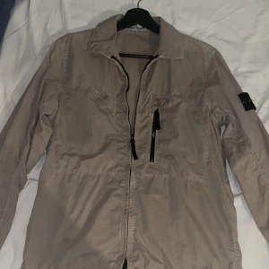 Beige Stone island jacka med dragkedja - Snygg beige jacka från Stone Island med dragkedja framtill och en praktisk ficka på bröstet. Jackan har långa ärmar och en klassisk krage. Perfekt för en stilren look. Knappt använd! pris kan diskuteras! Står ej för frakten, kan mötas upp i GBG