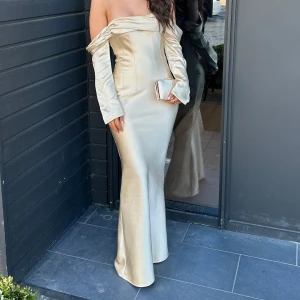 Champagnefärgad långklänning - Elegant champagnefärgad långklänning med offshoulder-design och långa ärmar. Klänningen har en figurnära passform som framhäver silhuetten.