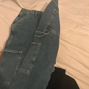 Blå cargopants i jeansmaterial - Säljer ett par blå cargopants i jeansmaterial med flera praktiska fickor. Byxorna har en loose passform och är perfekta för en avslappnad stil. De har en klassisk midja och stängs med dragkedja och knapp.