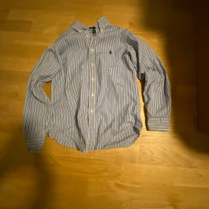 Blå och vit randig skjorta från Ralph Lauren - Säljer en klassisk blå och vit randig skjorta från Ralph Lauren. Skjortan har långa ärmar och knappar framtill. Den är broderad med den ikoniska logotypen på bröstet. Perfekt för en stilren look.