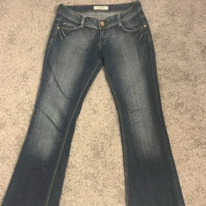 Mörkblå lågmidjade bootcut jeans med dekorerade bakfickor - Snygga lågmidjade mörkblå bootcut jeans med coola dekorationer på bakfickorna. De har en klassisk femficksdesign och en knappgylf. Perfekta för en avslappnad stil med en touch av bling. Aldrig andvönt köpt begagnat säljer då dom är för stora storlek 33 en knapp saknas bak