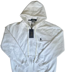 Vit hoodie från Polo Ralph Lauren - Säljer en stilren vit hoodie från Polo Ralph Lauren med dragkedja och klassisk logga på bröstet. Perfekt för en avslappnad look. Den har en justerbar huva och ribbade muddar vid ärmslut och nederkant. Jag fick den av en kompis så kan ej garantera om den är äkta. Jag skulle tro det för jag har kollat och gämnfört loggan, men jag kan inte garantera.