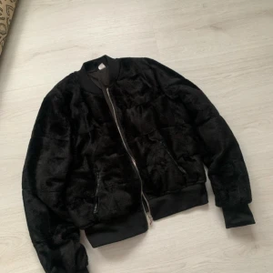 Svart pälsjacka med dragkedja - Snygg svart pälsjacka med dragkedja framtill. Jackan har en klassisk bomberjacka-stil med ribbade muddar vid ärmslut och nederkant. Perfekt för att hålla sig varm och stilren under kyligare dagar.