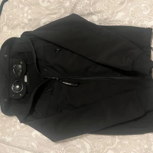 Svart windbreaker med glasögondetalj från C.P. Company - Säljer en svart windbreaker från C.P. Company med unika glasögondetaljer på huvan. Jackan har dragkedja framtill och flera praktiska fickor. Perfekt för dig som söker jacka till våren men håller även bra i kylan 