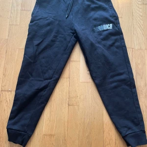 Svarta mjukisbyxor från Hoodrich - Säljer ett par svarta mjukisbyxor från Hoodrich med snörning i midjan och ribbade muddar vid bensluten. Byxorna har en tryckt logga på ena benet och är perfekta för en avslappnad stil.