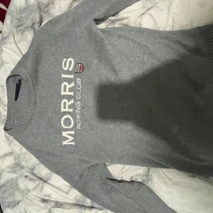 Grå tröja från Morris - Säljer en stilren grå tröja från Morris med texten 'Rowing Club' och en liten flagga på bröstet. Tröjan har långa ärmar och ribbade muddar vid ärmslut och nederkant. Perfekt för en avslappnad stil.