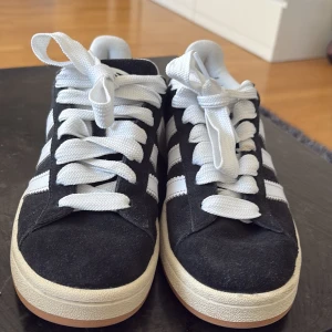 Svarta Adidas Campus 00s sneakers - Snygga svarta Adidas Campus 00s sneakers med vita detaljer och klassiska tre ränder. Skorna har en bekväm passform och är perfekta för en avslappnad stil. De är tillverkade i mocka och har snörning framtill.