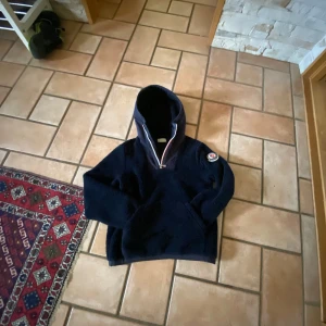 Svart hoodie från Moncler - Säljer en snygg svart cardigan från Moncler med en klassisk design. Tröjan har en huva och en dragkedja vid halsen. Perfekt för en stilren och bekväm look.