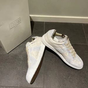 Masion Margela - Snygga vita sneakers från Maison Margiela med beige snörning, rund tå och en unik textur. Gummisula i brunt ger en snygg kontrast. Perfekta för en stilren look – och helt nya, aldrig använda. Pris går att snacka om. Skriv om du är nyfiken.