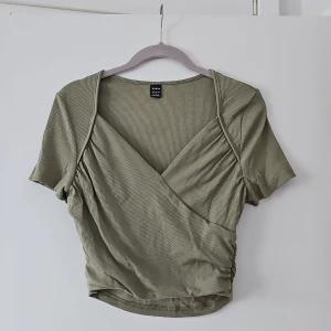 Olivgrön omlottopp från Shein - Säljer en olivgrön omlottopp från Shein med korta ärmar och ribbad struktur. Toppen har en smickrande omlottdesign och är perfekt för en stilren look. Passar bra till både jeans och kjol.