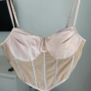 Beige korsettopp med dragkedja - Snygg beige korsettopp med smala axelband och dragkedja i ryggen. Toppen har dekorativa ränder med glittriga detaljer som ger en extra touch. Perfekt för att lyfta vilken outfit som helst!