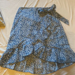 Blå blommig omlottkjol - Charmig blå omlottkjol med vita blommor och volangdetaljer. Kjolen har ett knytband i midjan för en justerbar passform. Perfekt för en somrig look!