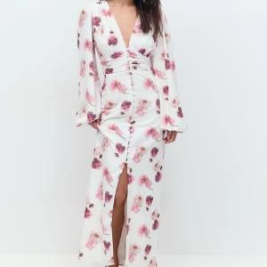 Vit blommig långklänning - Elegant vit långklänning med rosa blommönster och djup v-ringning. Klänningen har långa ärmar och en slits framtill som ger en stilfull touch. Perfekt för sommarens festligheter.