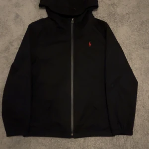Svart jacka med dragkedja från Ralph Lauren - Säljer en stilren svart jacka från Ralph Lauren med dragkedja och huva. Jackan har en liten röd logga på bröstet och långa ärmar. Perfekt för en casual look.