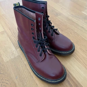 1460 Vinröda Dr. Martens - Snygga vinröda boots från Dr. Martens i skinn med klassisk snörning och gul söm runt sulan. Perfekta för en cool och stilren look. De har en robust design och är kända för sin hållbarhet.