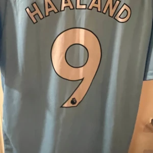 Manchester City fotbollströja - Snygg ljusblå Manchester City fotbollströja och shorts  med HAALAND och nummer 9 på ryggen. Tröjan har klubbens emblem på bröstet och Etihad Airways som sponsorlogga. Perfekt för fans av laget!