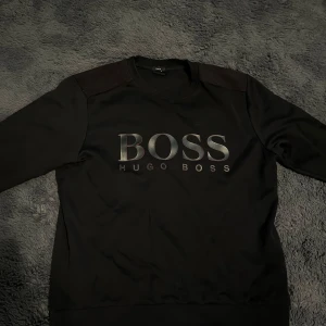 Hugo Boss tröja - Säljer en stilren svart tröja från Hugo Boss med deras logga i svart med rött runt omkring på framsidan. Tröjan har en slim fit passform och långa ärmar, med svart röd sydda axlar.