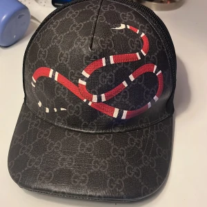 Svart keps med ormtryck - Snygg svart keps från Gucci med ett iögonfallande rött ormtryck. Kepsen har ett diskret mönster och justerbar rem baktill. Perfekt för att ge din outfit en lyxig touch. Box och kvitto ingår.