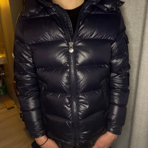 Mörkblå Moncler Maya - Tja! Säljer en snygg mörkblå Maya från Moncler. Jackan har en glansig finish och är utrustad med en dragkedja framtill. Otroligt fint skick ingen defekt eller liknande! Den har långa ärmar och en huva för extra värme. Perfekt för kyliga dagar! Givetvis äkta! Vid fundering är den endast att höra av sig!