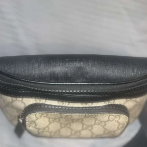 Beige och svart väska från Gucci - Snygg beige och svart väska från Gucci med klassiskt mönster. Väskan har en dragkedja och justerbart bälte för enkel användning. Perfekt för att bära dina nödvändigheter med stil.