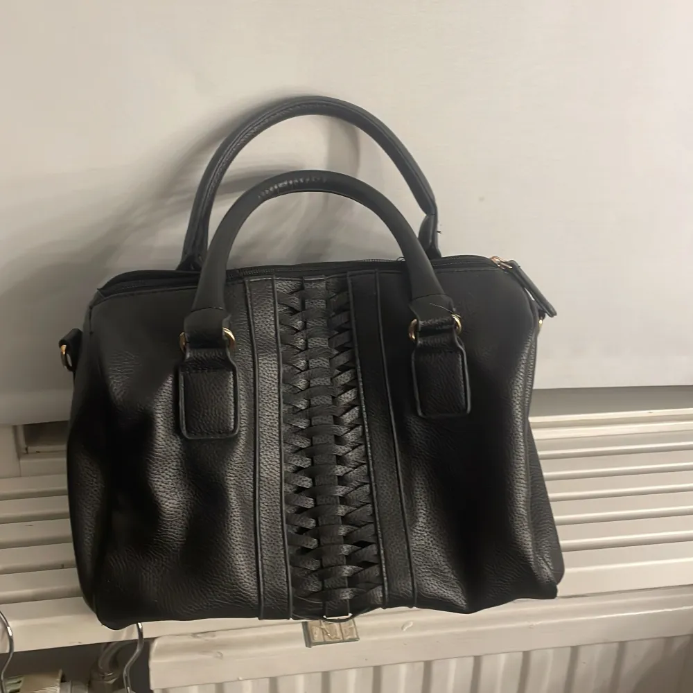 Snygg svart handväska från Atmosphere med flätad detalj på framsidan. Väskan har två handtag och en dragkedja upptill. Perfekt för att bära dina dagliga nödvändigheter med stil.. Laukut & Käsilaukut.