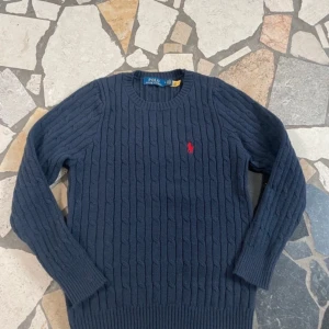 Mörkblå stickad tröja från Ralph Lauren - Säljer en snygg mörkblå stickad tröja från Ralph Lauren med det klassiska röda logotypbroderiet på bröstet. Tröjan har en rund halsringning och långa ärmar. Perfekt för kyligare dagar!