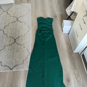 Grön axelbandslös långklänning - Elegant grön axelbandslös långklänning i ett slätt material. Perfekt för en festlig kväll med sin stilrena design och långa fall. Klänningen har en smickrande passform som framhäver figuren. Även slits